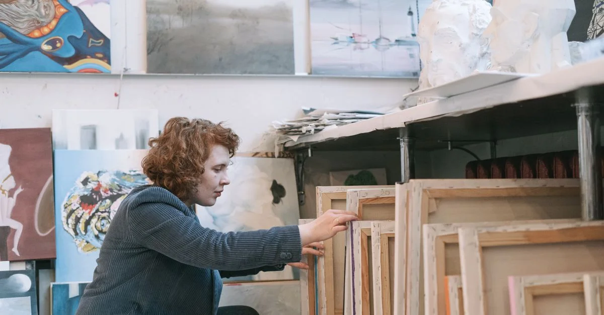Une artiste concentrée organise des toiles dans un studio d'art en désordre rempli d'œuvres d'art diversifiées.