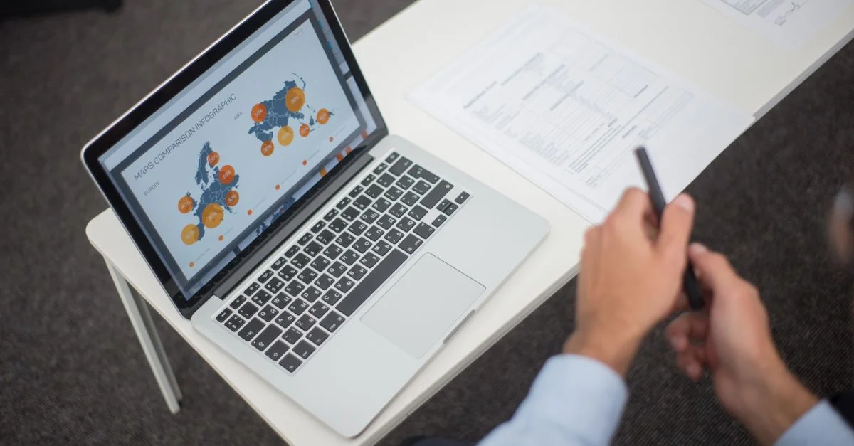Professionnel analysant des infographies de données sur un ordinateur portable à un bureau, avec des documents et un stylo.