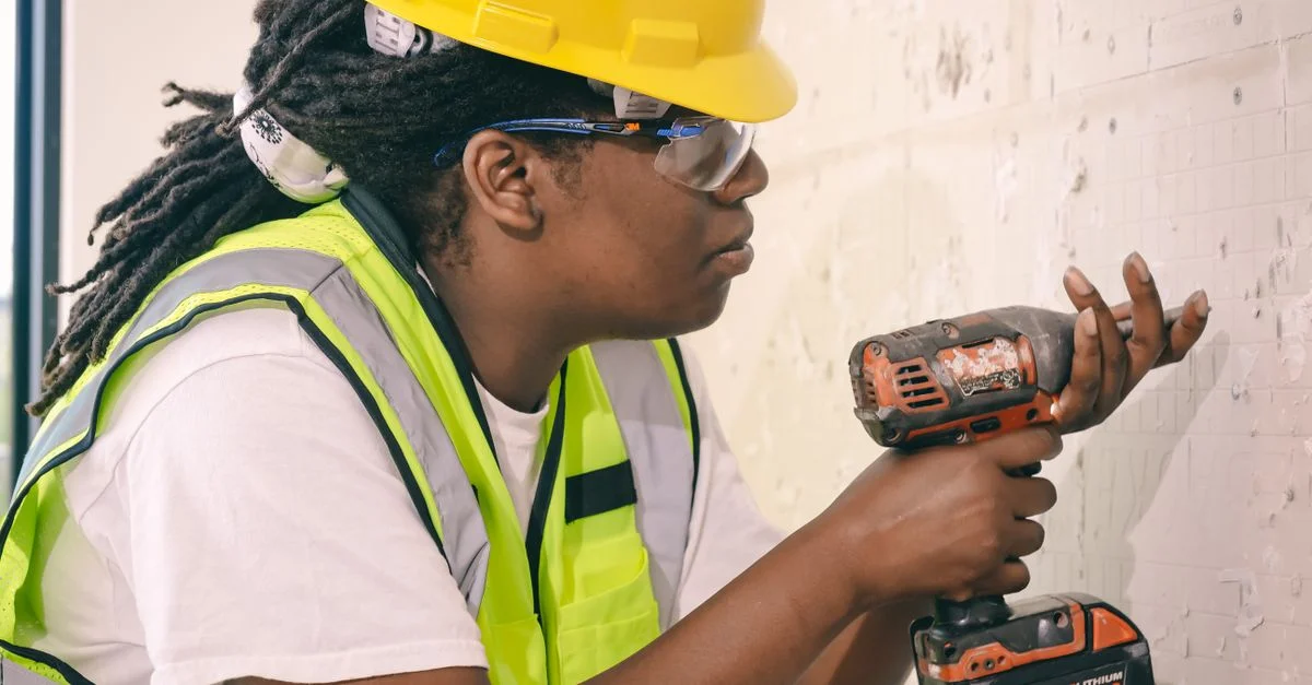 Une ouvrière du bâtiment utilisant une perceuse, portant des équipements de sécurité à l'intérieur.