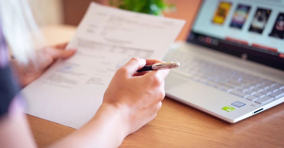 Gros plan sur une main tenant un stylo et du papier près d'un ordinateur portable, suggérant des achats en ligne ou la révision de documents.