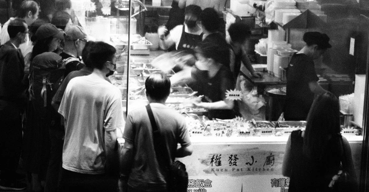 Image en noir et blanc d'un stand de street food très fréquenté à Hong Kong, avec des gens qui font la queue.