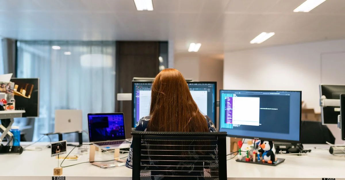 Vue arrière d'une ingénieure logiciel travaillant sur un poste de travail à plusieurs moniteurs dans un bureau.
