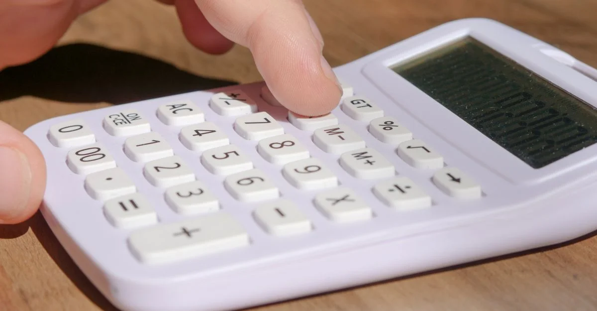 Une main appuyant sur les touches d'une calculatrice blanche posée sur un bureau en bois sous une lumière vive.