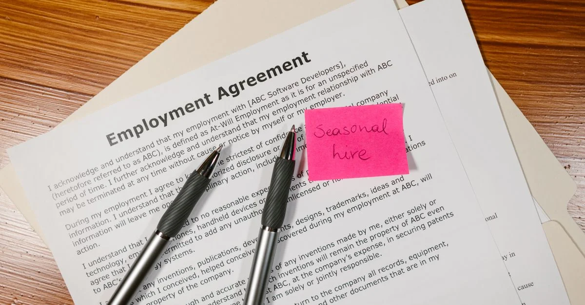 Vue de dessus d'un contrat de travail avec des stylos et une note de recrutement saisonnier sur un bureau en bois.