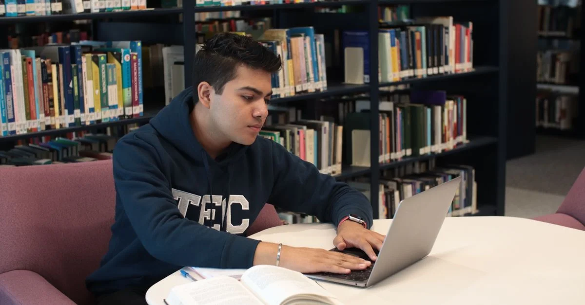 Étudiant dans une bibliothèque utilisant un ordinateur portable et des manuels pour étudier et faire des recherches.