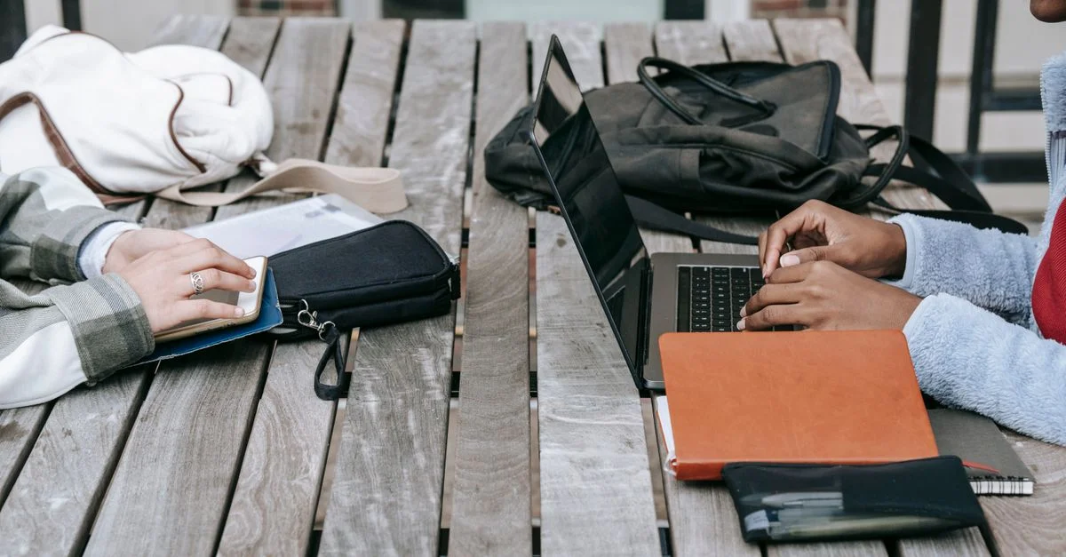 Étudiantes travaillant sur un projet en plein air, utilisant des ordinateurs portables et des carnets sur une table en bois.